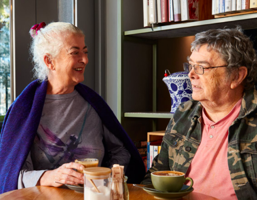 Two older women sit at a table with coffee, having a yarn.